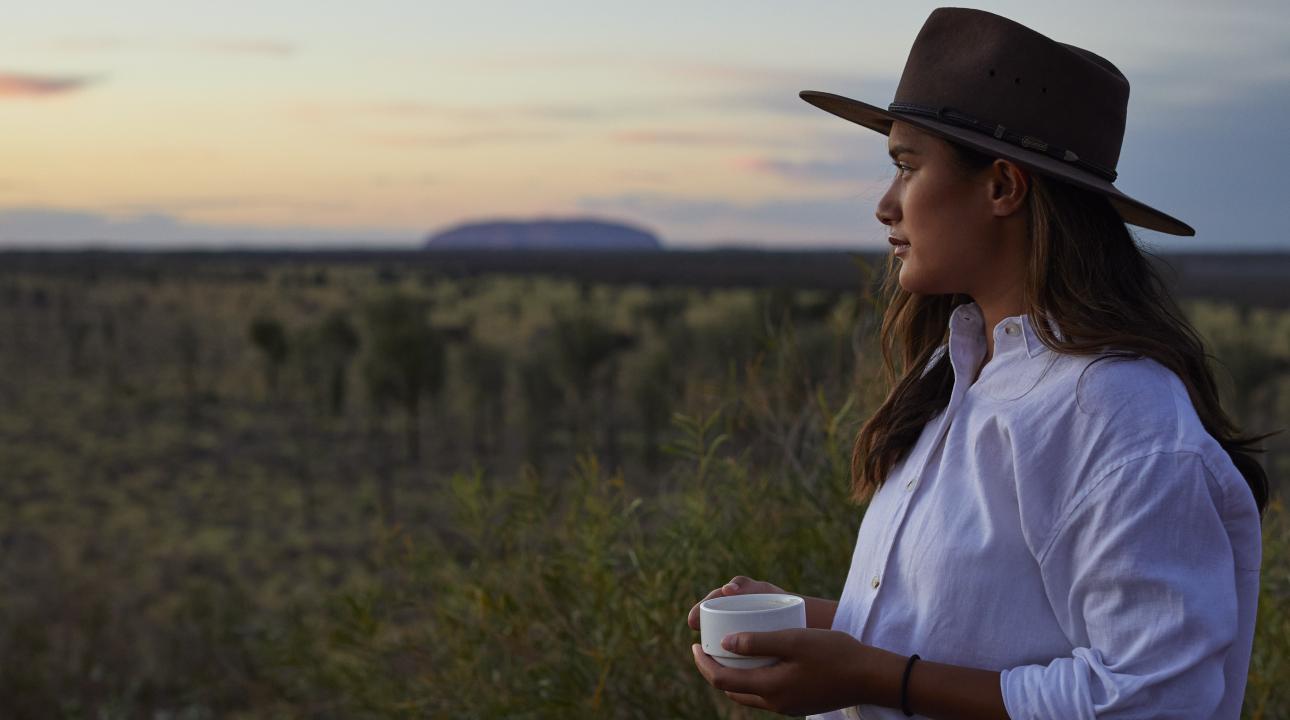 Woman drinking coffee staring into the sunrise with Uluru Rock in the distance