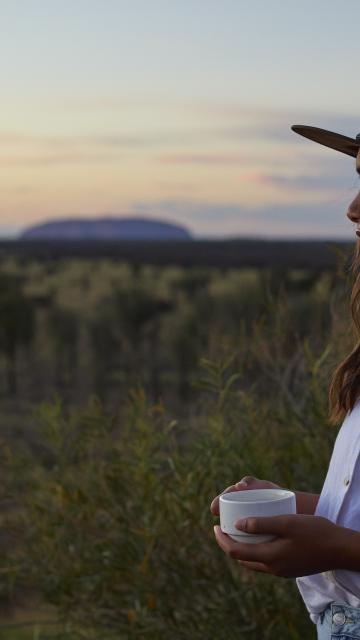 Woman drinking coffee staring into the sunrise with Uluru Rock in the distance
